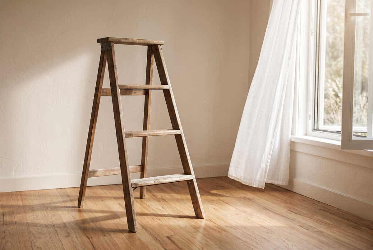 A short wooden step ladder standing in a sunlit room beside an open window, the bottom rung worn smooth from use